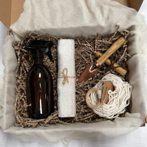 Photo of products inside a large gift box. An amber glass bottle with black atomiser spray, a natural micro fibre cloth, a cream macrame plant hanger, with a set of copper mini gardening tools sit in a bed of recycled paper stuffing on muslin fabric.