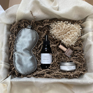 Photo of products inside the new parent gift box. Grey sleep mask, amber glass bottle of room spray and silver comfort balm pot, cotton body buffer and pot of lip balm sit on a bed of recycled brown paper stuffing sitting on organic muslin fabric.