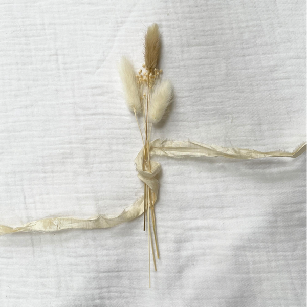 Small set of white and natural brown bunny tail dried grass cuttings laid centrally on a white muslin fabric background. The small bundle has a natural sari silk ribbon woven around it, across the composition of the photo. 