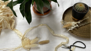 Aerial flatlay on a white muslin fabric background. A bundle of natural sari silk ribbon, dried flowers, with bonsai shears are laid out across the frame. Leaves from a plant appear top centre, with a gold tray holding a cactus and dried flowers right.