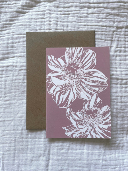 Pink greetings card with a white line illustration of an anemone flower layed ontop of a kraft paper A5 envelop. Both sit on a muslin fabric background.