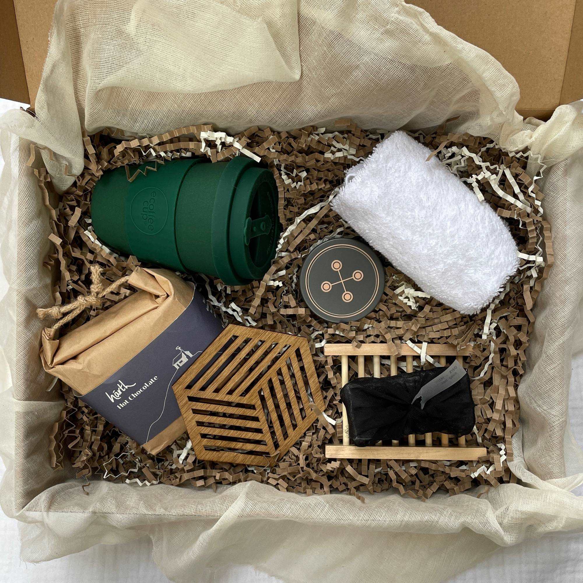 Photo of products inside a cardboard gift box. A deep evergreen eCoffee Cup, 1 geometric hexagonal birchwood coaster, a rectangular brown paper package of hot chocolate, white face cloth, small tin of XO Balm, black soap bar and bamboo tray sit on recycled brown paper stuffing inside organic muslin fabric.