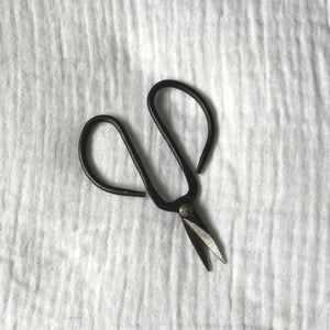 A pair of metal mini bonsai pruning shears lay open pointing downwards on white cotton muslin fabric.