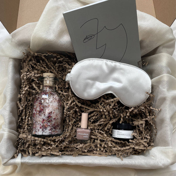 Photo of products inside a carboard gift box. A grey notebook, white silk sleep mask, clear glass bottle of rose bath salts, nude nail varnish and face mask pot sit on a bed of recycled brown paper stuffing on organic muslin fabric.