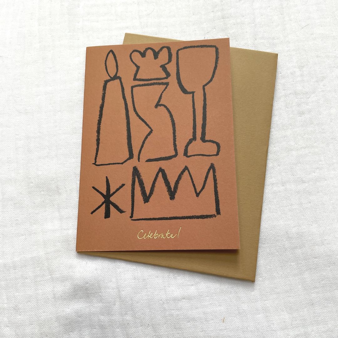 Photo of a burnt orange card and gold envelop laid on a white cotton muslin fabric. The card has minimalist abstract shapes on it with gold embossed writing that says 'Celebrate'.