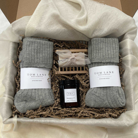 Photo of products inside a large cardboard gift box. Two grey pairs of Tom Lane alpaca bed socks, blush pink soap bar sitting on a wooden soap slat dish and amber glass medium candle on a bed of recycled brown paper stuffing on organic muslin fabric.