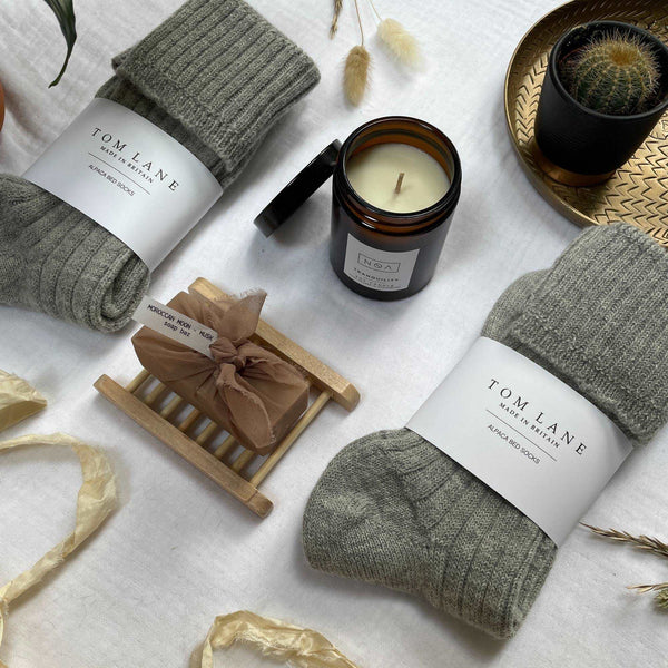 Contents of the large cosy gift box laid out on cotton muslin. Two pairs of grey bed socks, a candle and vegan soap bar on a bamboo rack. Recycled silk yarn lays to the bottom of the image, with dried flower heads and leaves to a plant appearing from the left top corner. A small cactus is on a gold tray top right.