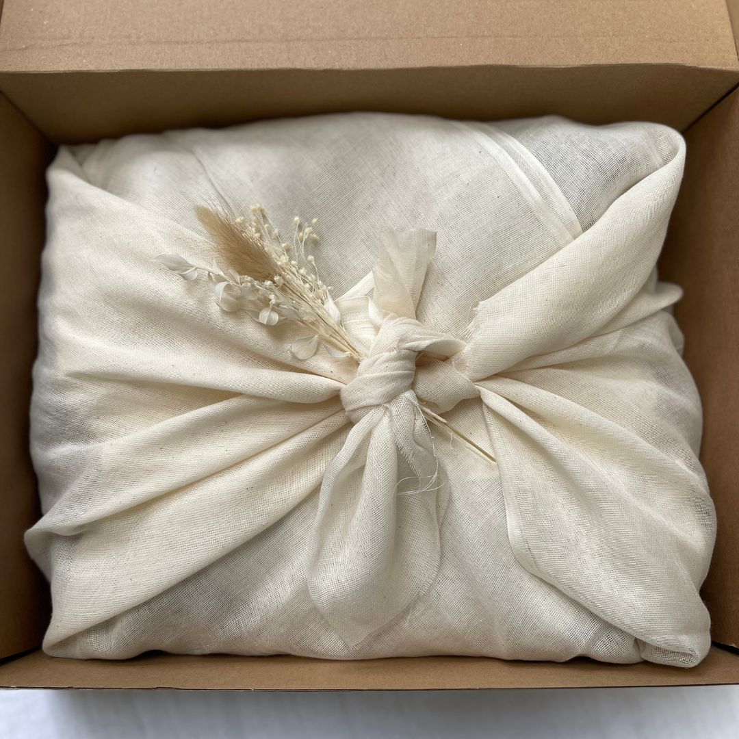 Aerial photo of the inside of a cardboard box. Contents wrapped in natural muslin fabric tied using the furoshiki method in the centre of the box. A small bunch of natural dried flowers appear placed within the tied fabric in the centre.