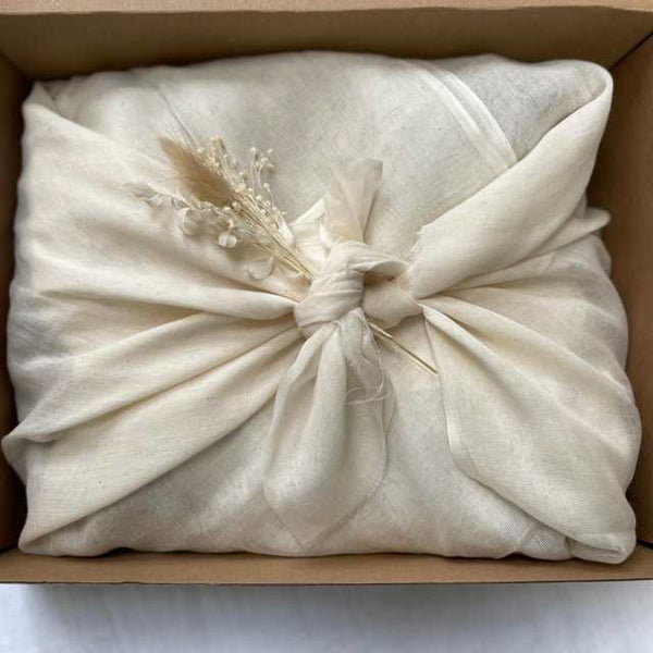 Aerial photo of the inside of a cardboard box. Contents wrapped in natural muslin fabric tied using the furoshiki method in the centre of the box. A small bunch of natural dried flowers appear placed within the tied fabric in the centre.