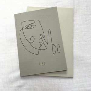 Photo of a grey card and cream envelop laid on a white cotton muslin fabric. The card has minimalist abstract line drawing of a face on it with gold foil writing that says 'Hey'.