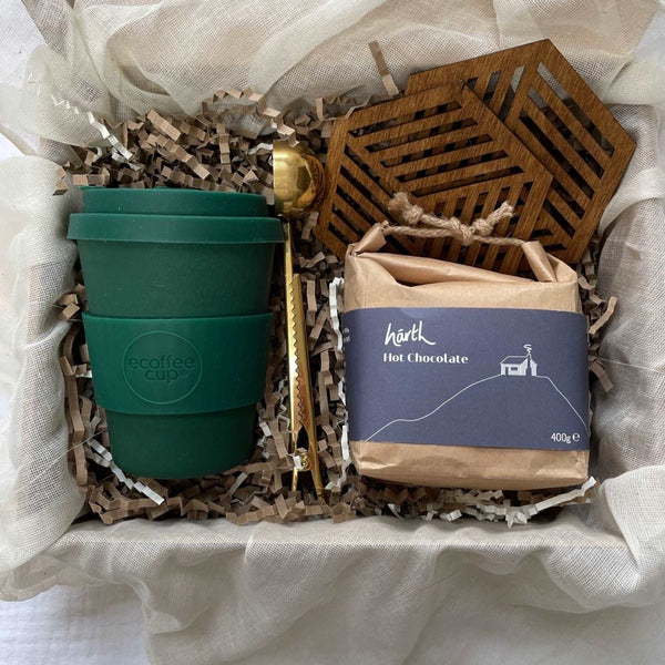 Photo of products inside a cardboard gift box. A deep evergreen eCoffee Cup, 2 geometric hexagonal beech wood coasters, a gold stainless steel coffee scoop and a rectangular brown paper package of hot chocolate sit on recycled brown paper stuffing inside organic muslin fabric.