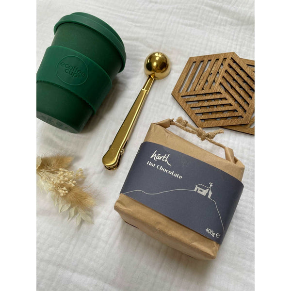 Contents of the hot bev lover gift box laid out on cotton muslin. 2 birchwood geometric drink coasters, an evergreen keep cup, gold stainless steel scoop and paper pouch of hot chocolate powder. 