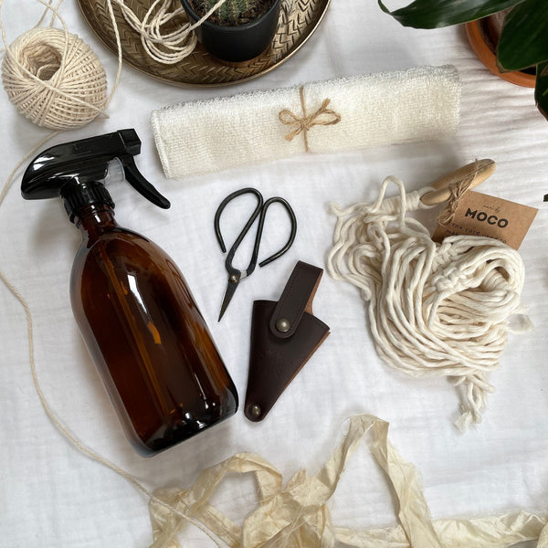 Aerial photo of an amber glass bottle with black atomiser spray head, a natural micro fibre cloth, a cream macrame plant hanger sits bundled in a ball along with a pair of metal bonsai shears and brown leather pouch. Silk sari yarn, dried flowers & plants surround the items.