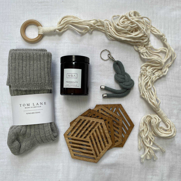 Contents of a large gift box laid out on cotton muslin. Grey alpaca bed socks, cream macrame plant hanger, 4 birchwood drink coasters, amber glass candle & grey key ring. 