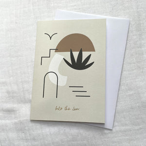 Photo of a cream card and white envelop laid on a white cotton muslin fabric. The card has minimalist abstract shapes on it with gold embossed writing that says 'Into the Sun'.