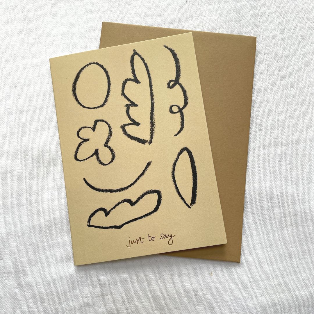 Photo of a burnt yellow card and gold envelop laid on a white cotton muslin fabric. The card has minimalist abstract shapes on it with gold embossed writing that says 'Just to Say'.