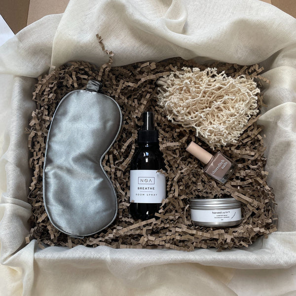 Photo of products inside a large cardboard gift box. Grey sleep mask, cream cotton body buffer, amber glass room spray bottom, nude nail varnish and silver comfort balm pot on a bed of recycled brown paper stuffing sitting on organic muslin fabric.