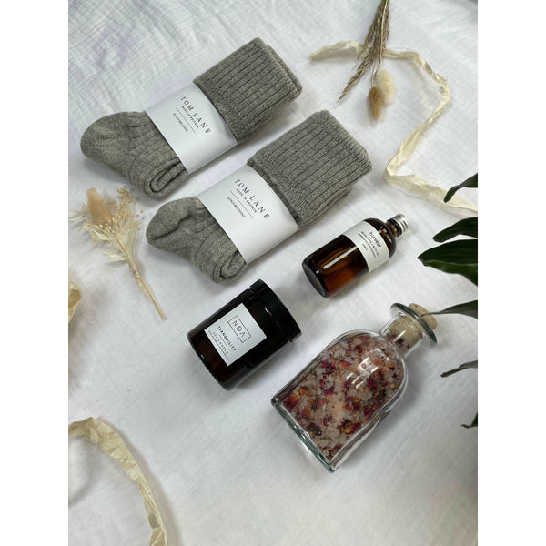 Photo of the items that feature in the newly weds gift box laid out on cotton muslin. A clear glass bottle of rose bath salts, two pairs of grey alpaca bed socks stacked to the right, an amber glass bottle of body oil and amber glass candle.