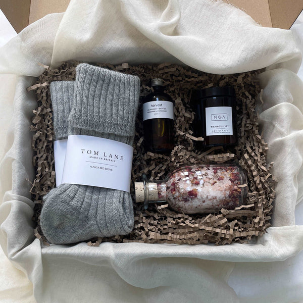 Photo of products inside a cardboard gift box. A clear glass bottle of rose bath salts, 2 pairs of grey alpaca bed socks, an amber glass bottle of body oil and amber glass candle sit on recycled brown paper stuffing on organic muslin fabric.