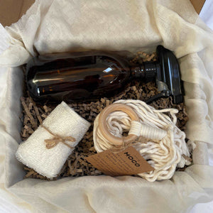 Photo of products inside a large cardboard gift box. An amber glass bottle with black atomiser, a natural micro fibre cloth, a cream macrame plant hanger sits bundled in a ball in a bed of recycled paper stuffing on muslin fabric.