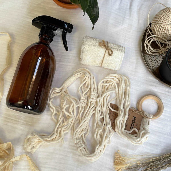 Aerial photo of an amber glass bottle with black atomiser spray head, a natural micro fibre cloth, a cream macrame plant hanger sits bundled in a ball. Silk sari yarn, dried flowers & plants surround the items