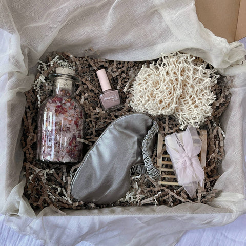 Photo of products inside a cardboard gift box. A grey silk sleep mask, clear glass bottle of rose bath salts, nude nail varnish, cotton body buffer and cream soap bar with bamboo tray, sit on recycled brown paper stuffing on organic muslin fabric.