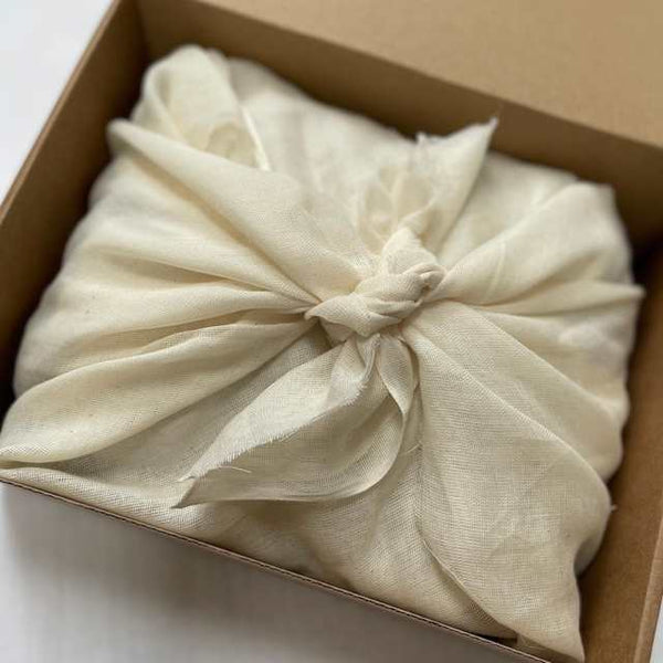 Aerial photo of the inside of a cardboard box. Contents wrapped in natural muslin fabric tied using the furoshiki method in the centre of the box.