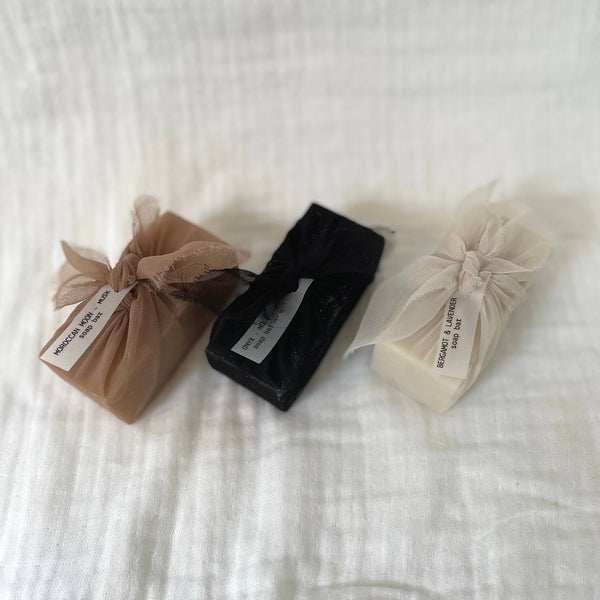 Photo of three bars of rectangular soap laid diagonally centred on a white muslin fabric background. They are each wrapped in sheer fabric using the furoshiki technique - blush, black and cream in colour. Each has a small white label tag attached.