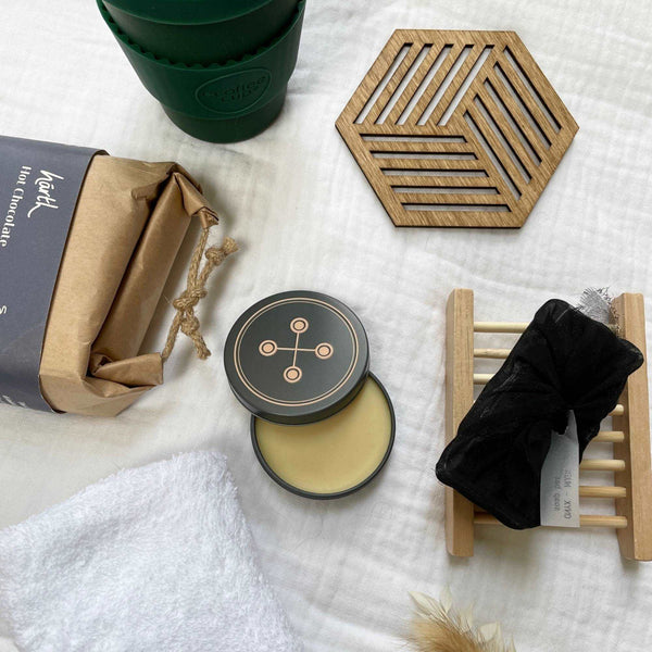 Aerial photo of a deep evergreen eCoffee Cup, 1 geometric hexagonal birchwood coaster, a rectangular brown paper package of hot chocolate, white face cloth, small tin of XO Balm, black soap bar and bamboo tray sit on white muslin fabric.