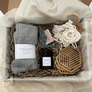Photo of products inside a large cardboard gift box. Grey alpaca bed socks, cream macrame plant hanger, 4 birchwood drink coasters, amber glass candle & grey key ring, sit in a bed of recycled brown paper stuffing on organic muslin fabric. 