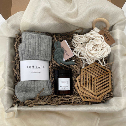Photo of products inside a large cardboard gift box. Grey alpaca bed socks, cream macrame plant hanger, 4 birchwood drink coasters, amber glass candle & grey key ring, sit in a bed of recycled brown paper stuffing on organic muslin fabric. 