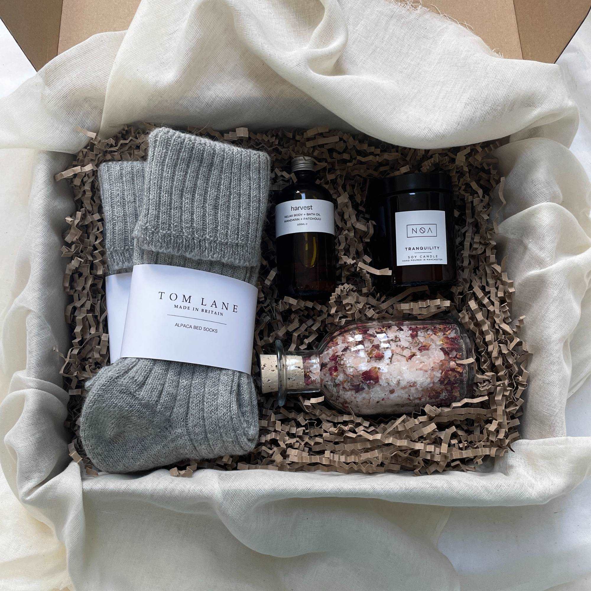 Photo of products inside a cardboard gift box. A clear glass bottle of rose bath salts, 2 pairs of grey alpaca bed socks, an amber glass bottle of body oil and amber glass candle sit on recycled brown paper stuffing on organic muslin fabric.