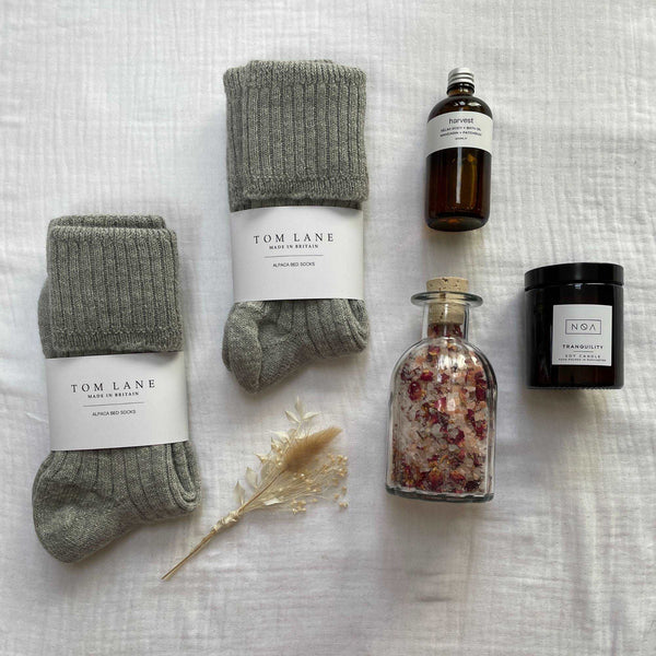 Photo of the items that feature in the wedding gift box laid out on cotton muslin. A clear glass bottle of rose bath salts, two pairs of grey alpaca bed socks stacked to the right, an amber glass bottle of body oil and amber glass candle.