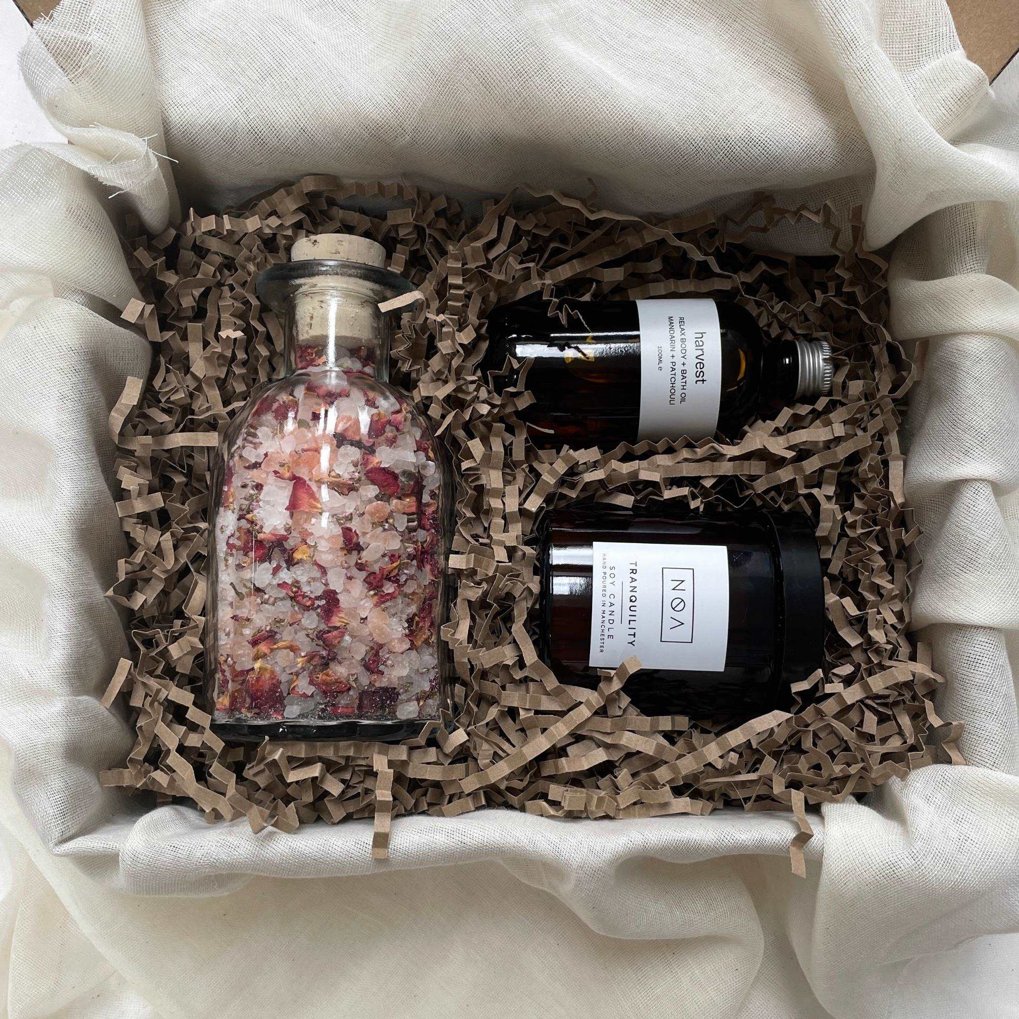 Photo of products inside a cardboard gift box. A clear glass bottle of rose bath salts, an amber glass bottle of body oil and amber glass candle sit on recycled brown paper stuffing on organic muslin fabric.
