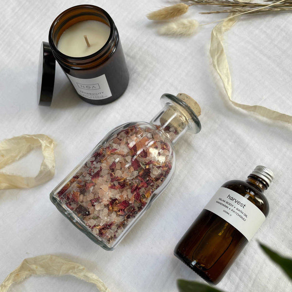 Photo of the items that feature in a gift box laid out on cotton muslin. A clear glass bottle of rose bath salts, an amber glass bottle of body oil and amber glass candle.