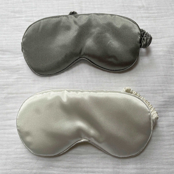 Photo of two silk face masks laid on a white fabric muslin background. Laid side by side the top mask is light grey with the bottom mask white in colour. Ruffled straps for each mask can be seen beneath appearing from the side of each face mask.