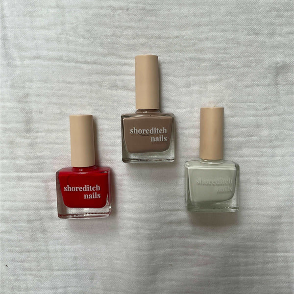 Photo of three nail varnishes laid centred on a white muslin fabric background. They are placed in an arrow formation of bright red, nude and white in colour.
